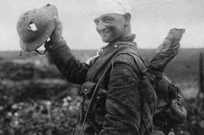 A soldier from a rare historical photo holding a damaged helmet and smiling on a battlefield.