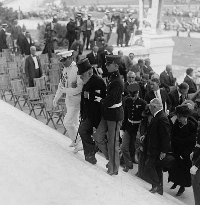 Rare historical photo of a man in a top hat being assisted up stairs by military personnel, with an audience observing.