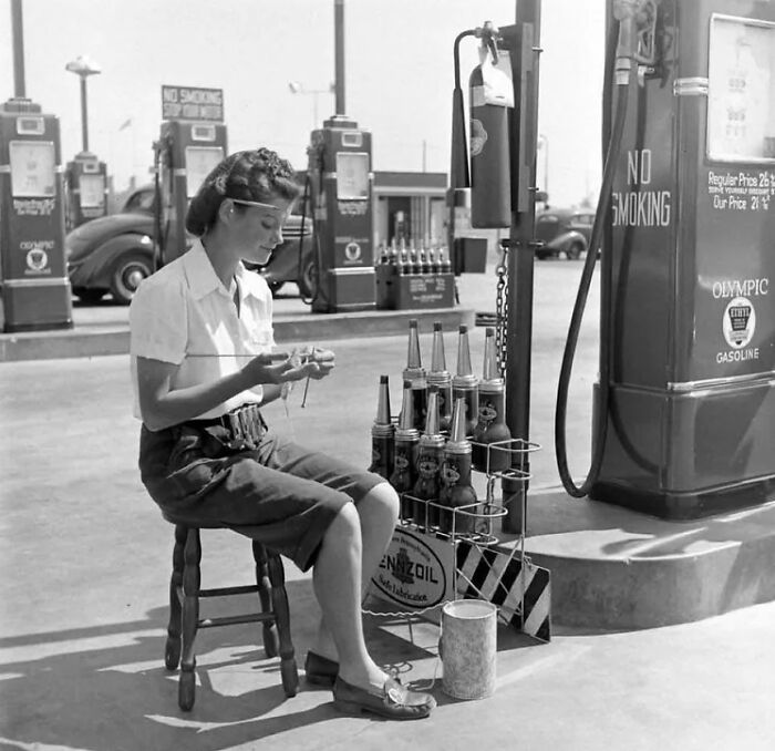 Woman knitting at an old gas station, surrounded by vintage oil cans and pumps, capturing a rare historical moment.