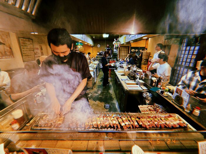 Man grilling skewers in a smoky Japanese restaurant, with diners in the background. Interesting Japan Pics atmosphere.