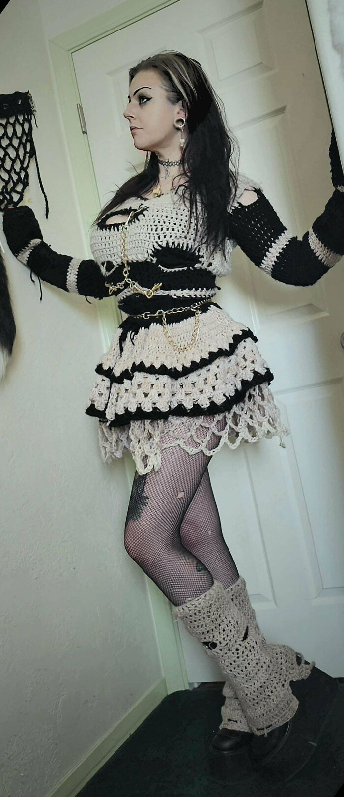 Woman wearing black and beige crochet dress and leg warmers paired with fishnet stockings and platform shoes indoors.