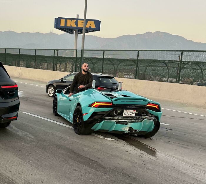 Man sits on a teal sports car with rear damage, parked on a highway, an example of a car accident and fail.