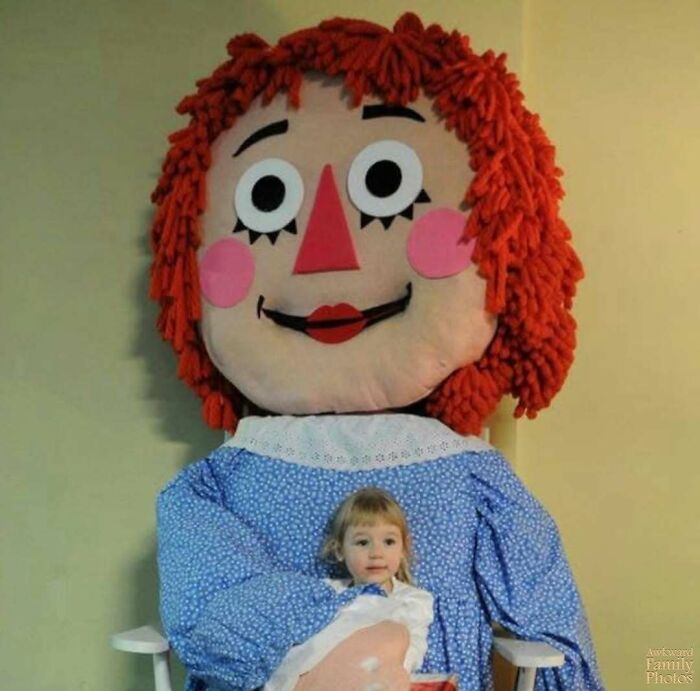 Child sitting on lap of an oversized doll with red yarn hair, capturing an awkward family photo with sweet vibes.