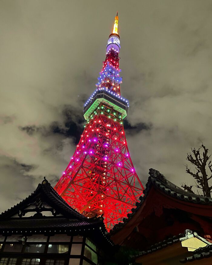 Tokyo Tower illuminated with colorful lights against a cloudy night sky, featuring traditional Japanese architecture in the foreground.