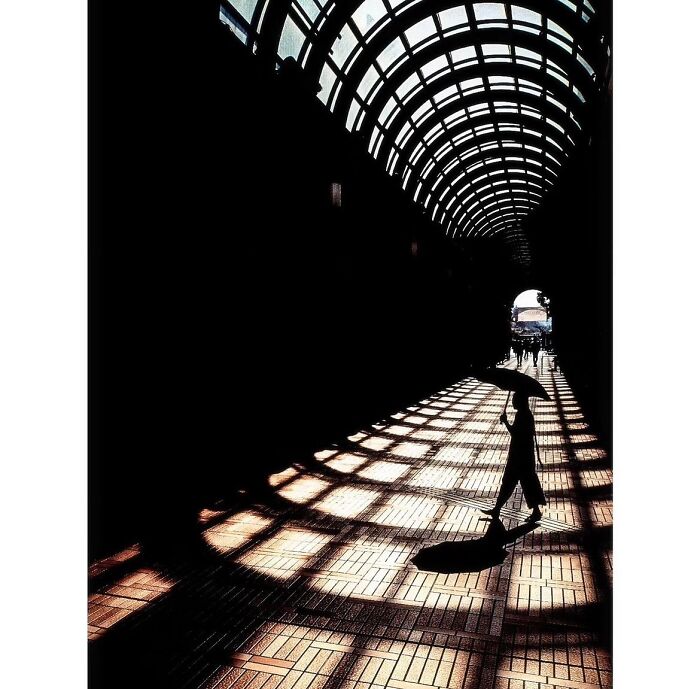Minimalist photo of a silhouette under an umbrella walking through a tunnel in Tokyo, casting geometric shadows.