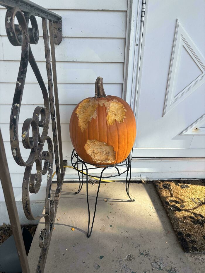 A "mildly interesting" pumpkin with bite marks, resembling a face, placed on a porch stand near a door.