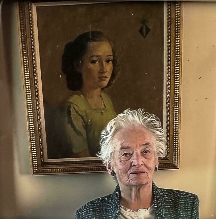 Elderly woman posing before a portrait of her younger self, highlighting the contrast between then and now.