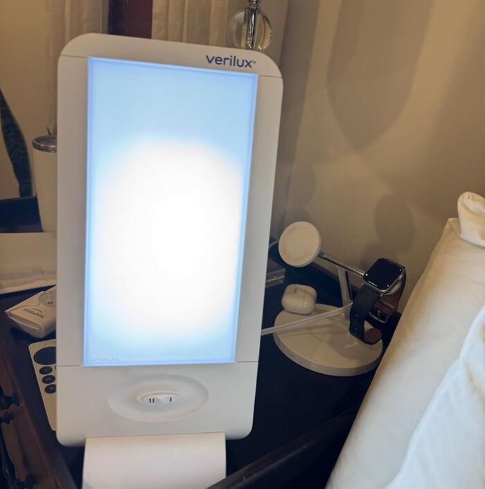 Verilux light therapy lamp on a desk with work from home tech essentials like smartwatch and earbuds.