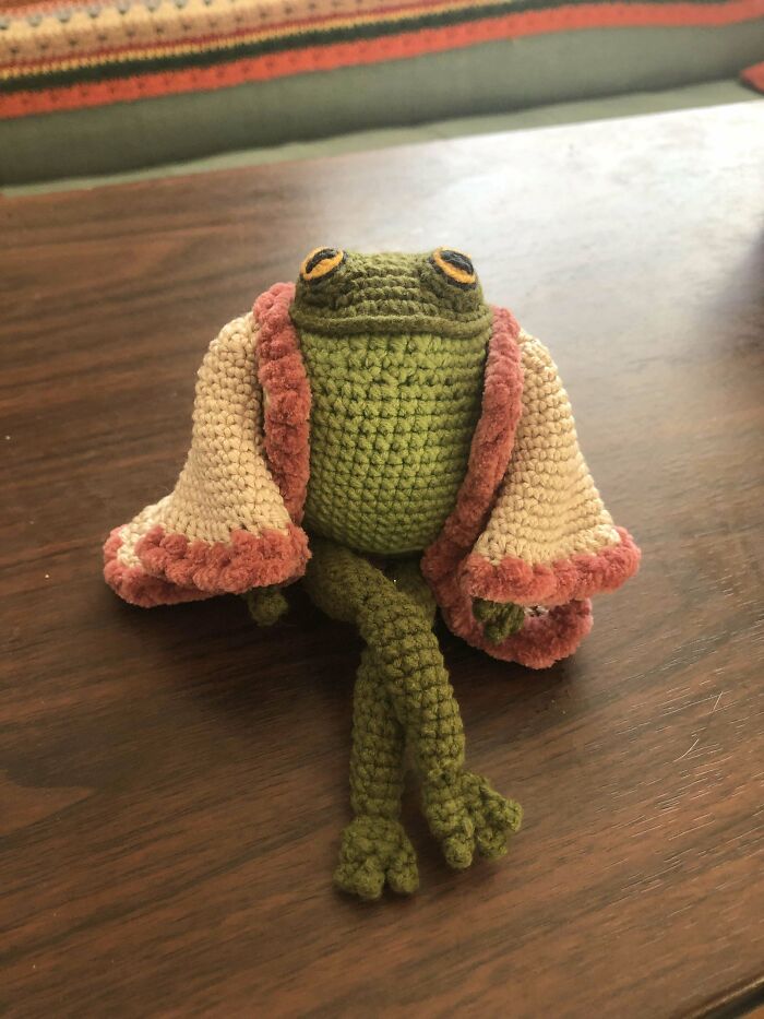 Crochet masterpiece of a relaxed green frog sitting with crossed legs, wearing a beige and rose-colored crocheted shawl.