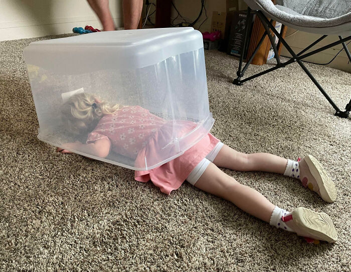 Niña haciendo tonterías divertidas, tumbada en el suelo con un contenedor transparente sobre ella.