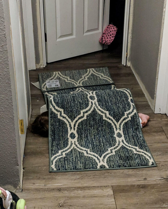 Child hiding poorly under a patterned rug in a hallway, showcasing a humorous take on cluelessness.