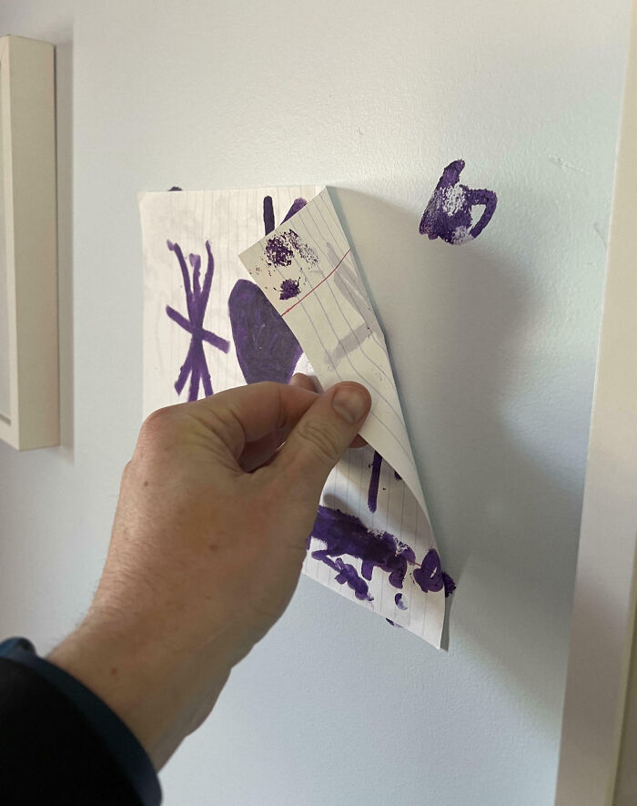 Hand peeling off child's artwork taped on a wall, revealing purple scribbles. Dumb-clueless-kids creativity moment.