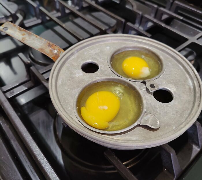 Old egg poacher on stove with two raw eggs in metal cups, showcasing cool-old-things.