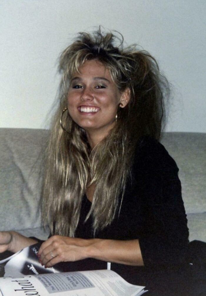 Woman with voluminous 80s-fashion hairstyle, wearing hoop earrings, reading a magazine and smiling.
