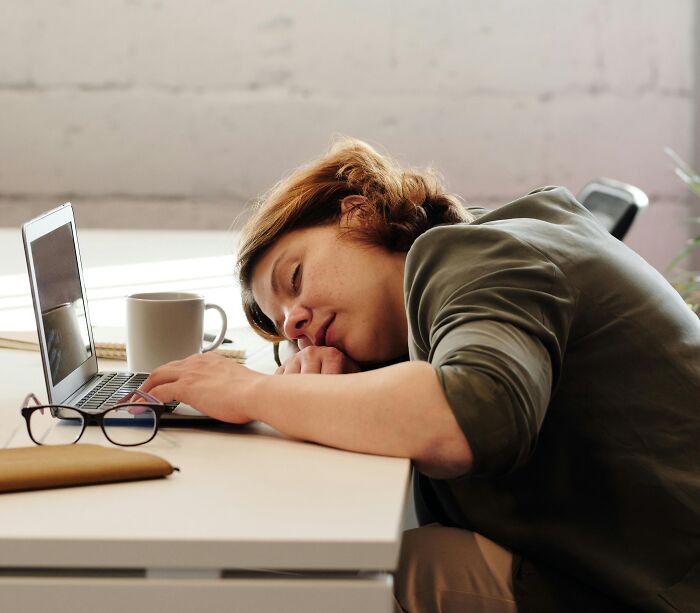 Person asleep on a desk in an office setting beside a laptop and coffee mug, illustrating a humorous office moment.