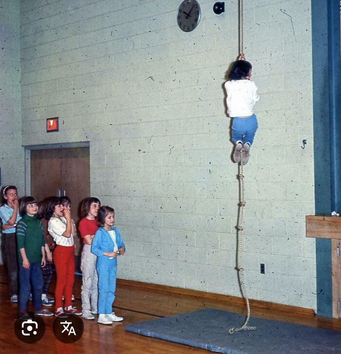 Children watching a girl climb a rope in a school gym, evoking old-school memories.