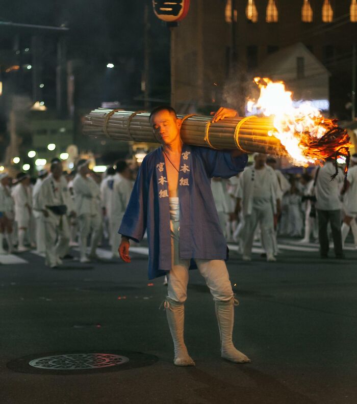 Man in traditional attire carrying a flaming torch, part of a cultural parade at night. Interesting-Japan-Pics.