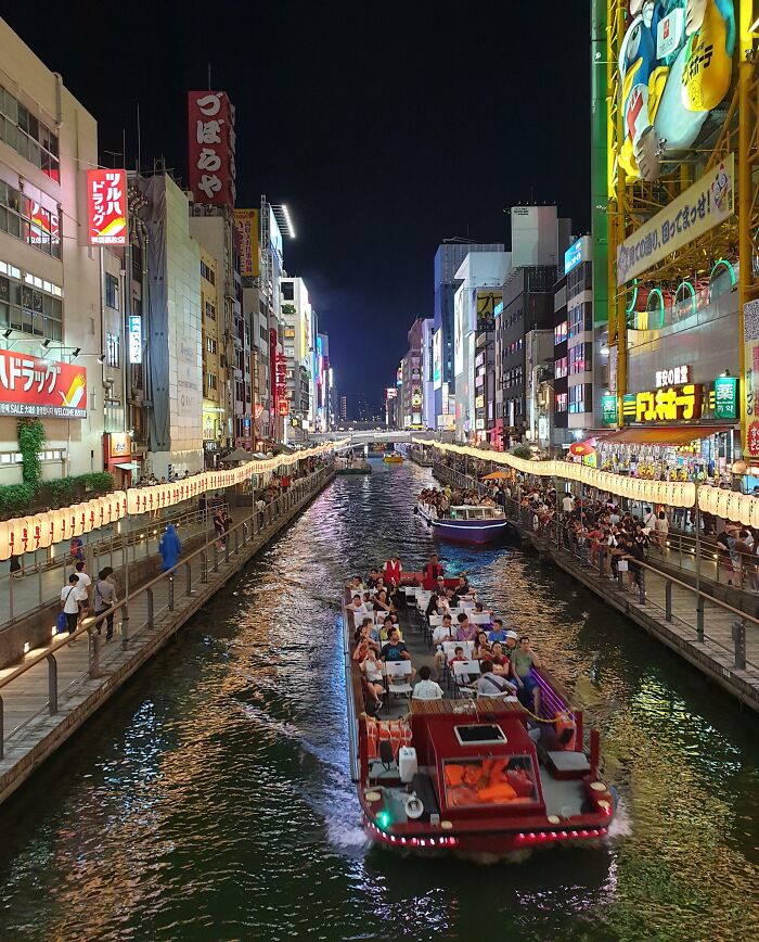 Vibrant night scene of a boat on a busy canal in Japan, surrounded by colorful city lights and crowds on the riverbanks.