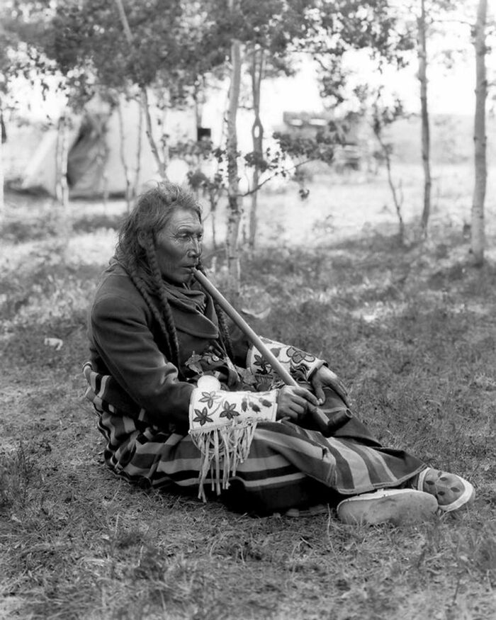 A historical image of a person sitting outdoors, wearing traditional attire and smoking a long pipe.