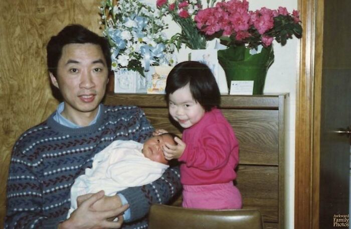 Man holding a newborn baby with a smiling young child beside him in an awkward family photo setting