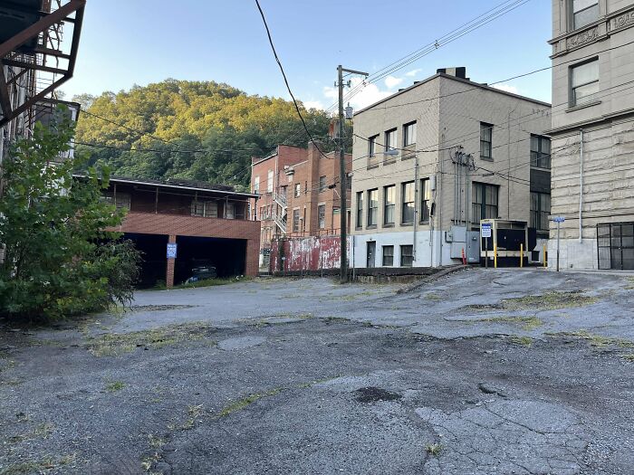 Urban decay scene with c*****d pavement, rundown buildings, and overgrown plants against a hilly backdrop.