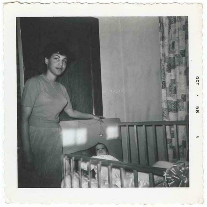 Woman standing beside a crib holding a toy above a baby in a black and white vintage photo from 1958 old images collection.