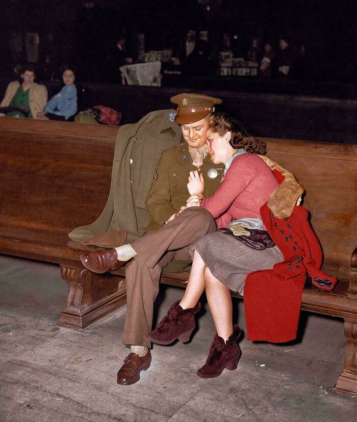 A soldier and woman sit closely on a bench in a historical setting, embracing lovingly.