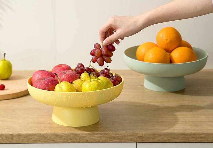 Hand picking grapes from a modern fruit bowl filled with apples and pears, next to another bowl of oranges on a wooden surface.