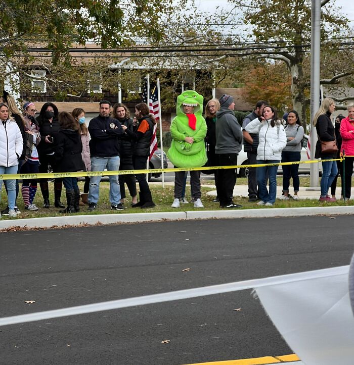 A person in a green dinosaur costume stands humorously among a crowd at an outdoor event, highlighting a hilarious misunderstanding.