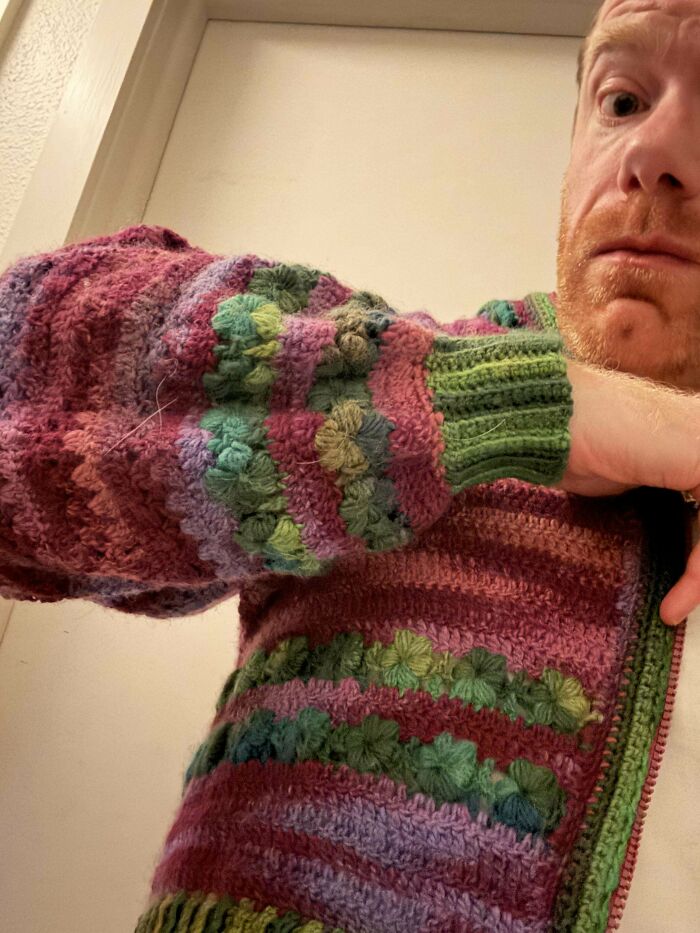 Man wearing a colorful handmade crochet sweater with textured floral patterns and green ribbed cuffs indoors.