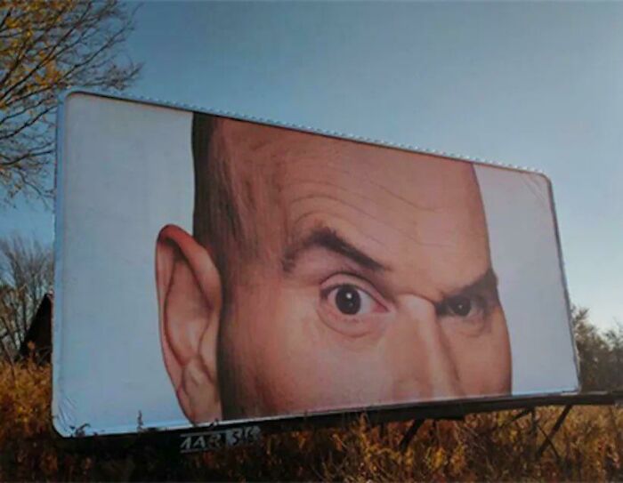 Funny billboard with a close-up of a man's face, showing a skeptical expression in a rural setting.