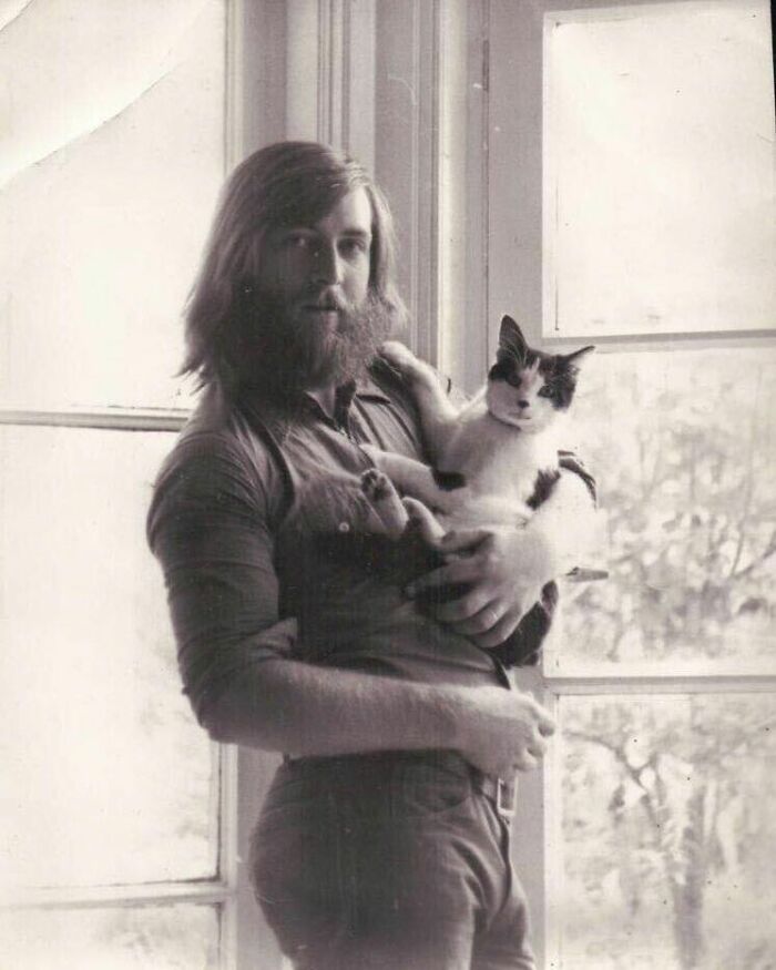 A man with a beard holding a cat by a window, in a black and white vintage photo.