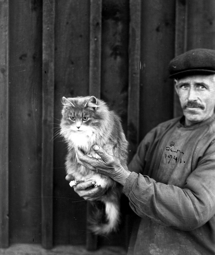 Man holding a fluffy cat in a vintage photo, wearing a hat and standing against a wooden background.