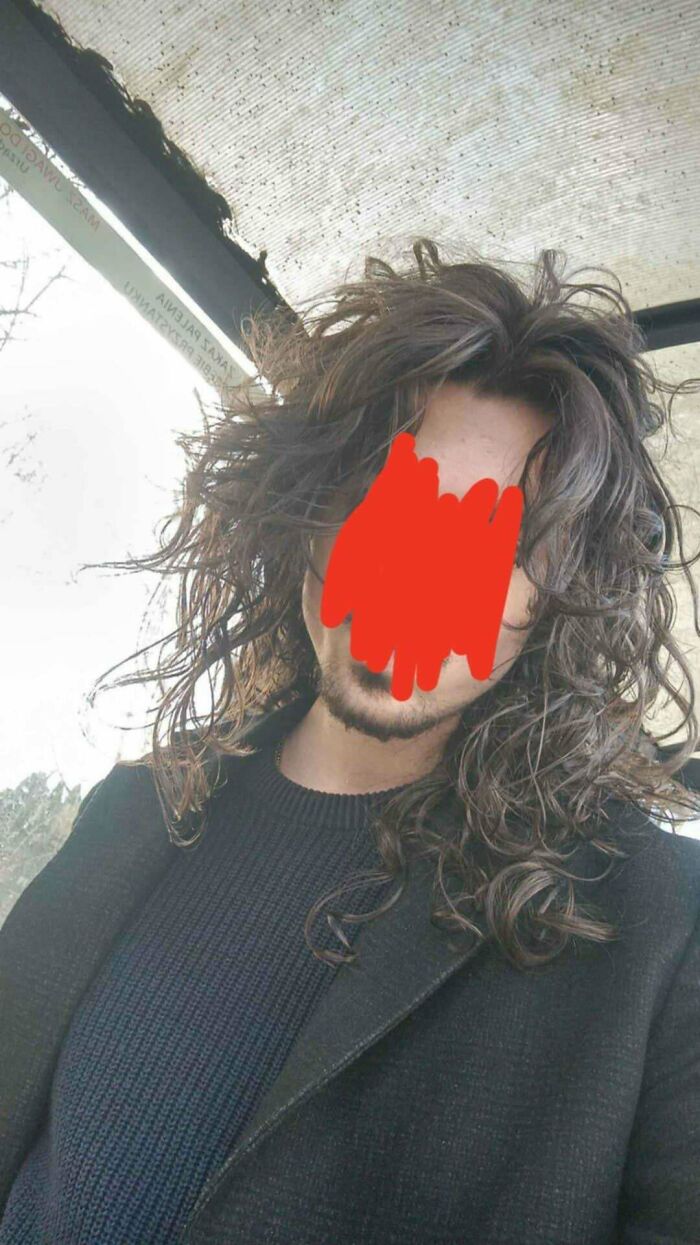 Man with fierceflow long curly hair wearing a black sweater and coat in an outdoor setting with a glass roof above.