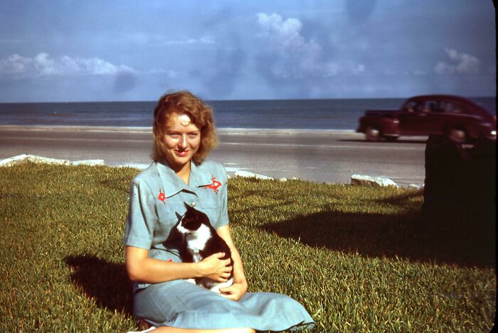 A woman in a vintage scene holding a cat by the seaside with a classic car in the background.