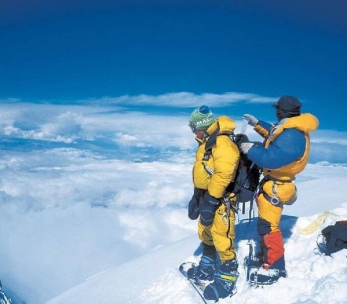 Mountaineers in yellow gear stand on a snow-covered peak, surrounded by a vast view of clouds and blue sky.