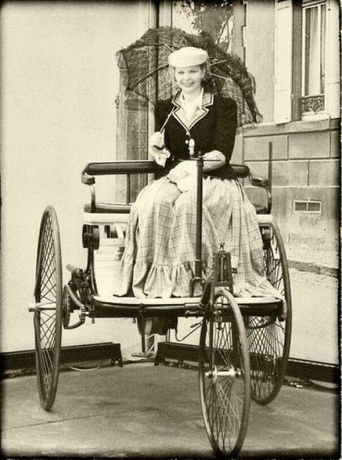 Woman in vintage attire smiles while sitting on an antique bicycle, illustrating an interesting historical fact.