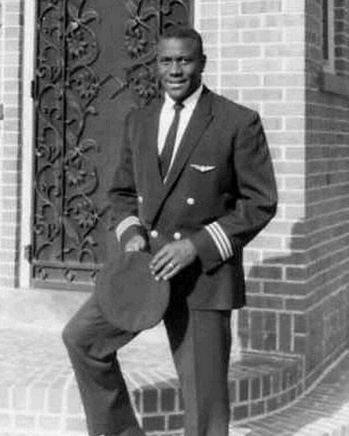 Black pilot in uniform poses in front of a brick building, highlighting an interesting fact shared by people.