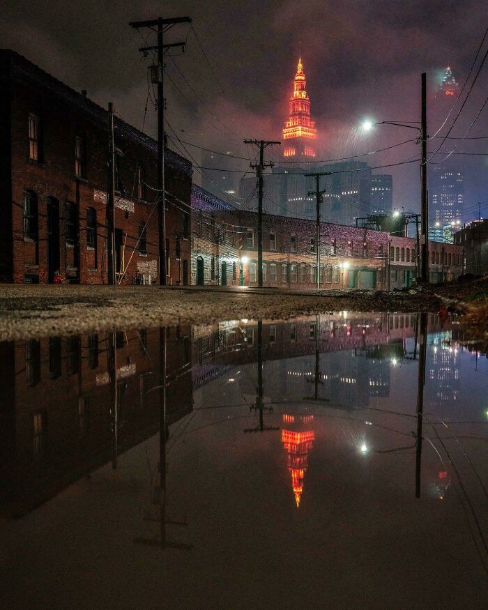 Dark urban street at night with old buildings and a glowing tower reflected in a large puddle, evoking supervillain headquarters vibes.