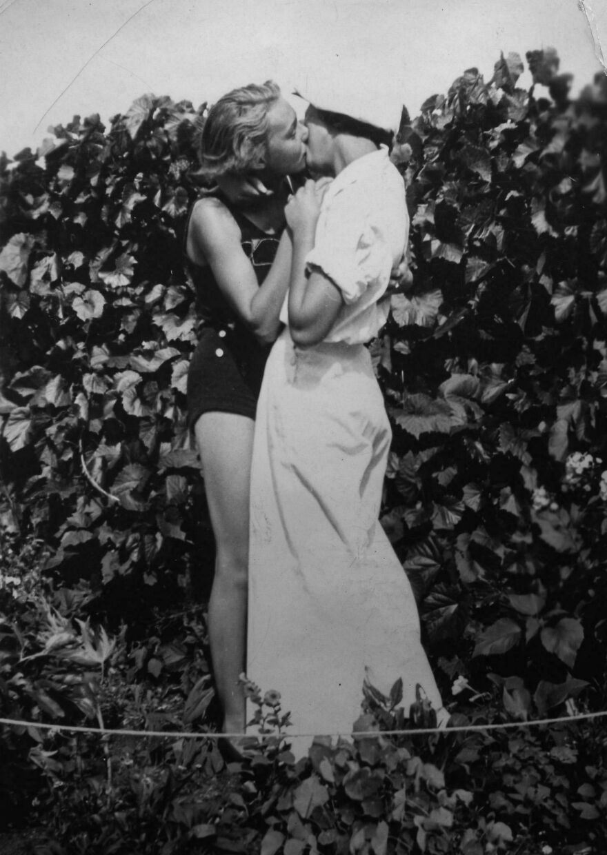 Historical photo of two people embracing and kissing in front of leafy bushes.