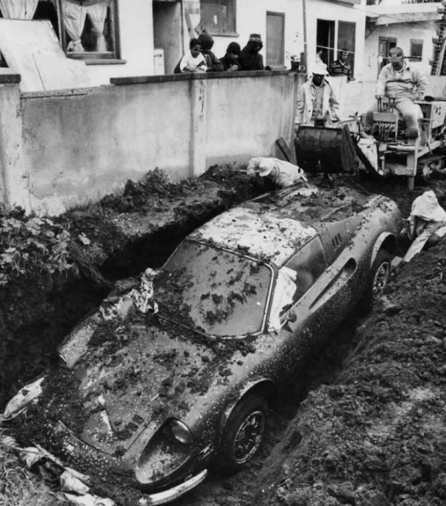 Historical photo of workers uncovering a buried vintage car in a residential area, with onlookers behind a wall.