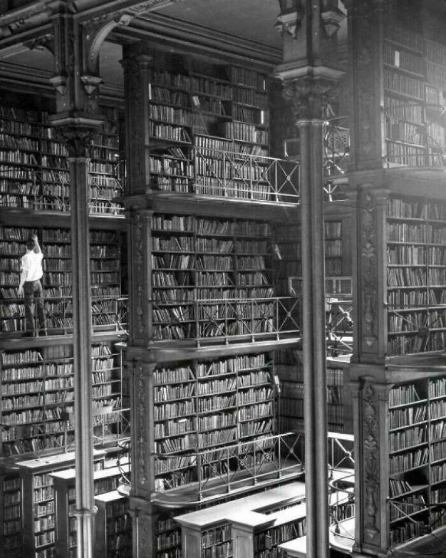 Historic photo of a man standing on a high library shelf, surrounded by countless books in a grand, multi-level library.