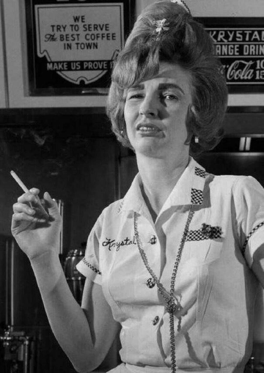 Woman in diner uniform holding a cigarette, standing by a sign about coffee.