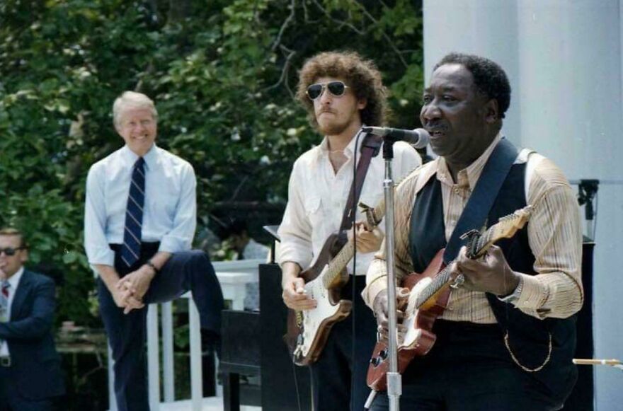 Historical photo of musicians performing outdoors, with a man in a suit watching nearby, capturing a vibrant moment.