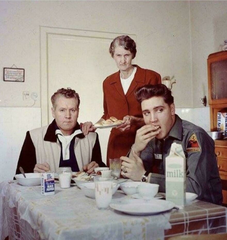A historical photo of three people at a kitchen table, with one person eating and another serving food.