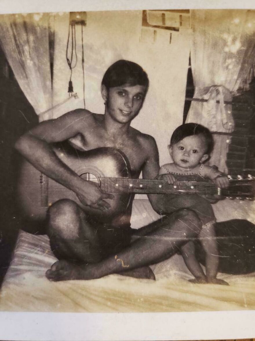 A historical photo of a shirtless man playing guitar next to a child sitting on a bed, with a vintage curtain backdrop.