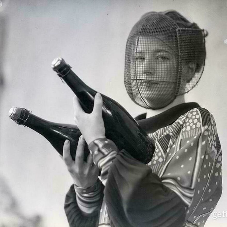 A woman in a vintage outfit holding large bottles, with a mesh face covering; historical photo.