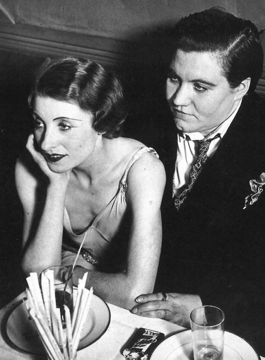 Two people seated at a table, one with hand resting on chin, in a vintage black-and-white historical photo.