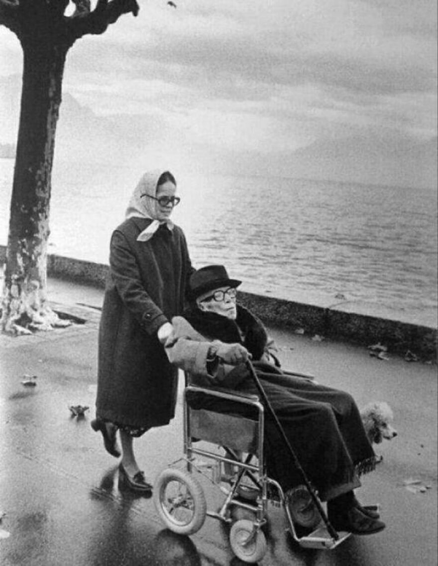 Two women in coats, one in a wheelchair, by a lake in a historical photo.