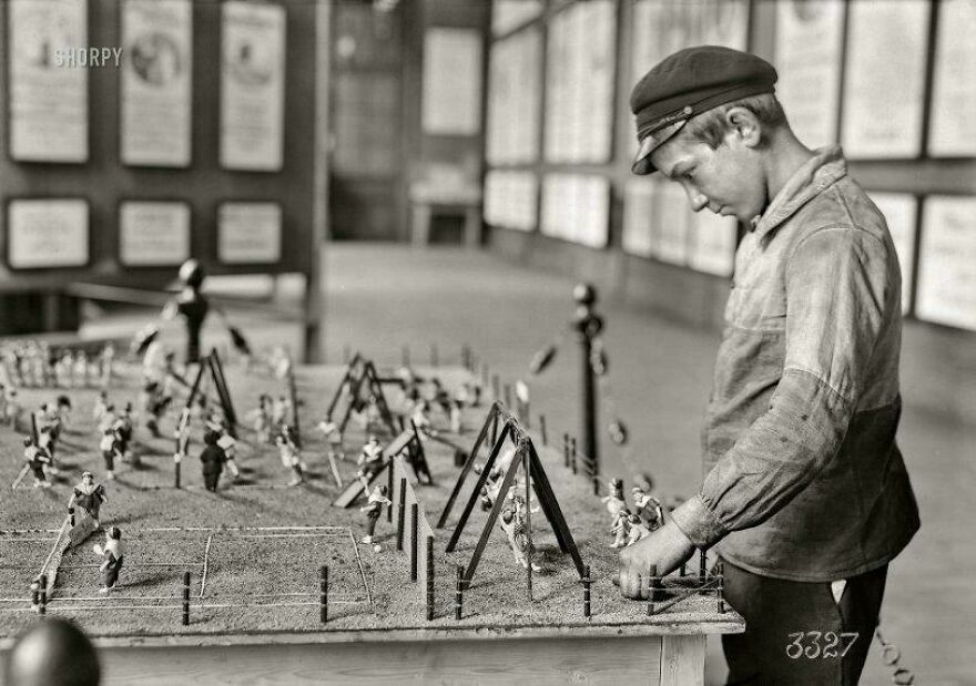 A historical photo of a boy playing with a vintage tabletop soccer game, wearing a cap and shirt in a museum setting.